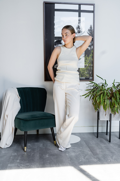 full length of woman in white posing near black armchair and looking away