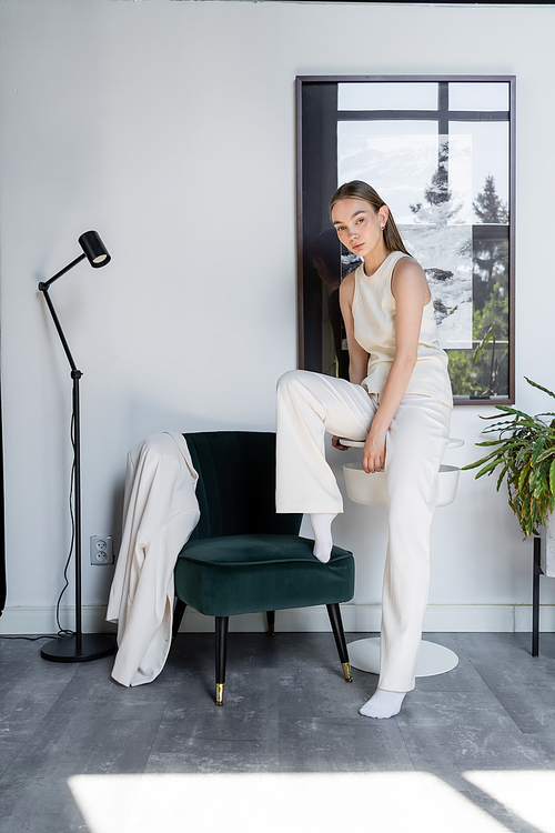 full length of woman in white trousers posing near black armchair and modern floor lamp