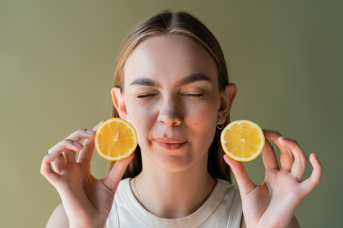 grimacing woman with closed eyes holding halves of sour lemon isolated on green