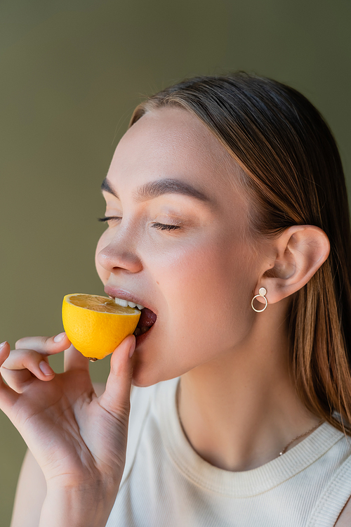 young woman with closed eyes biting juicy lemon isolated on green