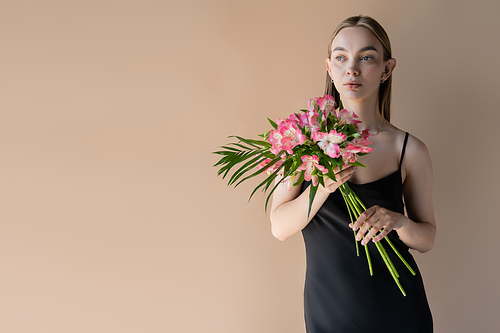 young woman in black strap dress holding bouquet of pink flowers isolated on beige