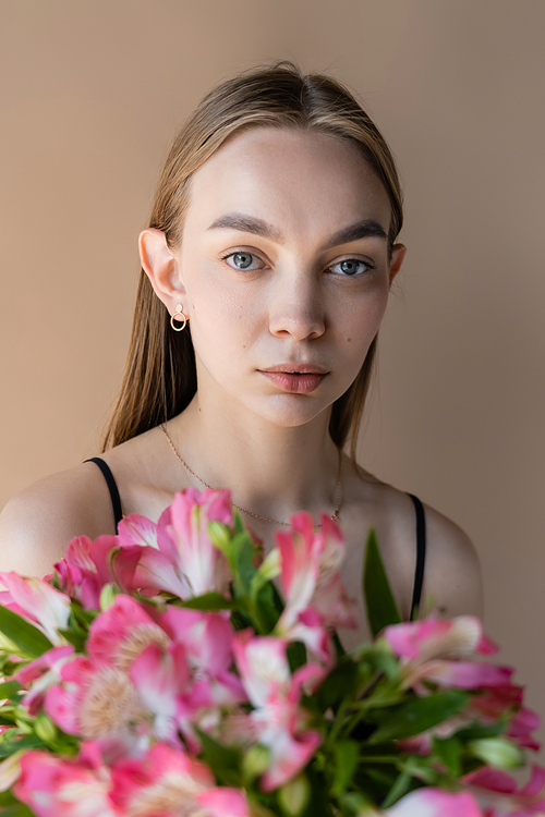 young woman with perfect skin looking at camera near alstroemeria flowers isolated on beige