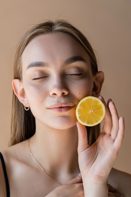 portrait of pleased woman with closed eyes holding half of juicy lemon isolated on beige