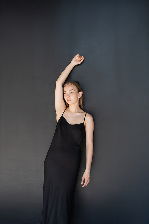 slim woman in strap dress posing with raised hand on black background