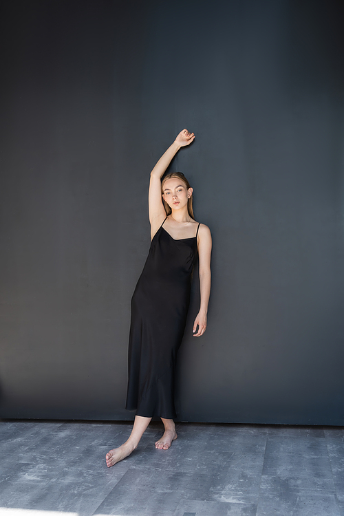 full length of barefoot woman in black strap dress posing with raised hand near dark wall