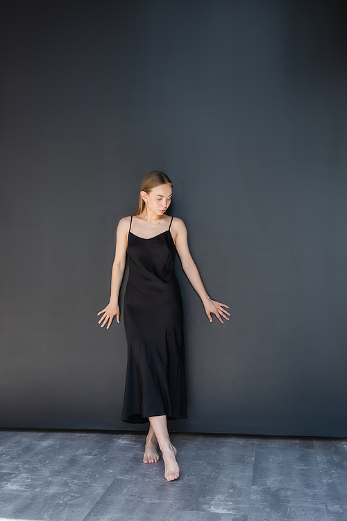 full length of young barefoot woman in strap dress standing near black wall