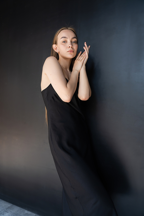 slim and seductive woman in black strap dress looking at camera near dark wall