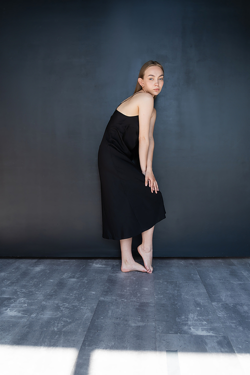 full length of slim barefoot woman in black strap dress near dark wall