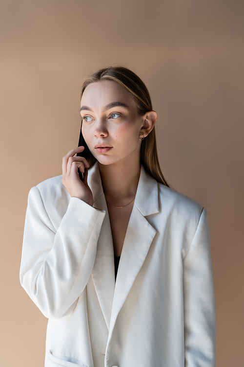 young woman in white blazer looking away while talking on smartphone isolated on beige
