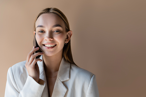 trendy woman smiling at camera while talking on mobile phone isolated on grey