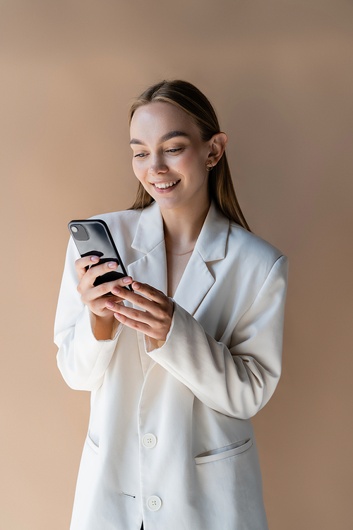 happy young woman in white blazer messaging on mobile phone isolated on beige