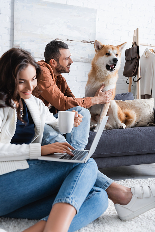 smiling young woman with cup using laptop near boyfriend cuddling akita inu dog