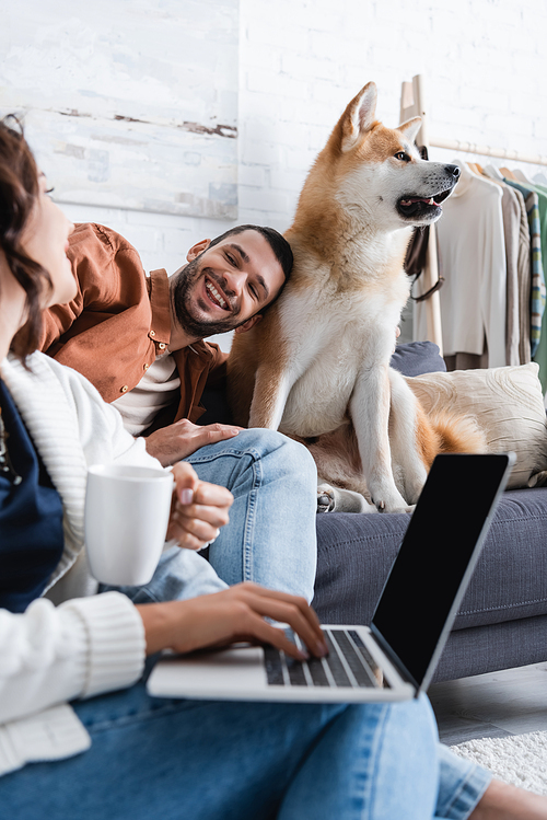 smiling young woman with cup using laptop near happy boyfriend leaning on akita inu dog