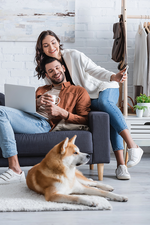 smiling young woman holding smartphone near boyfriend with laptop and looking at akita inu dog in living room