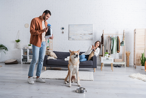 young woman sitting on couch and taking photo of boyfriend with pet food packaging and akita inu dog
