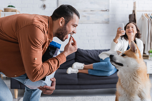 bearded man holding pet food near open mouth and looking at akita inu dog near blurred girlfriend with smartphone