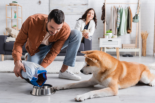 happy man adding pet food in bowl near akita inu dog and blurred girlfriend