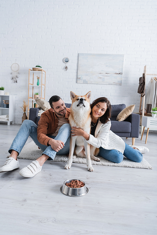 happy young couple hugging akita inu dog near bowl with pet food in living room