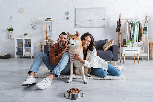 cheerful young couple hugging akita inu dog near bowl with pet food in living room