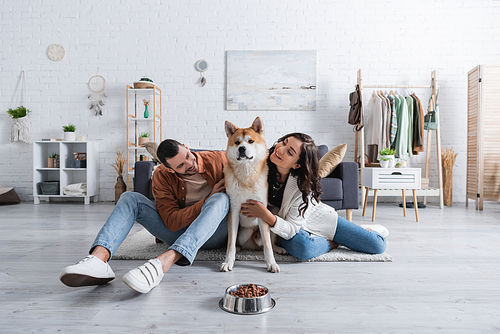 smiling young couple hugging akita inu dog near bowl with pet food in living room