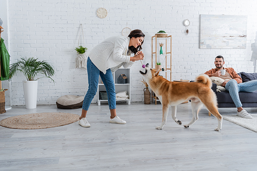 happy young woman playing with akita inu dog near bearded boyfriend with cup of coffee sitting on couch