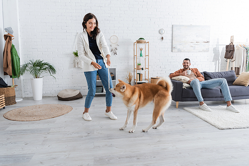 smiling young woman playing with akita inu dog near happy boyfriend with cup of coffee sitting on couch