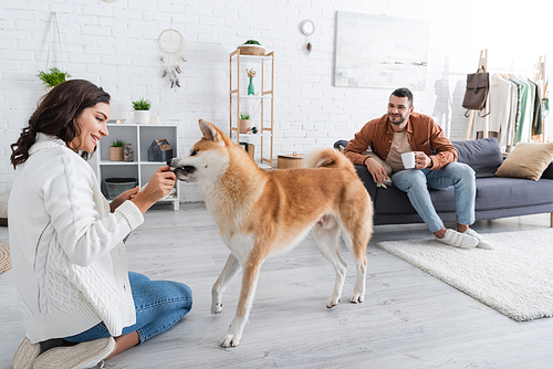 happy young woman playing with akita inu dog near bearded boyfriend with cup of coffee