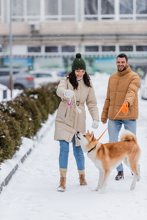 happy man holding leash while walking near cheerufl girlfriend playing with akita inu dog in winter