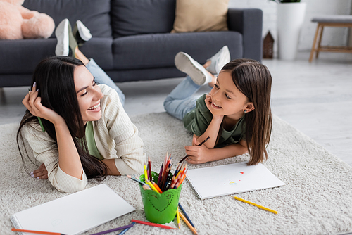 smiling nanny and girl looking at each other while lying on carpet near blank papers in living room