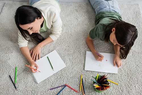 top view of nanny and girl drawing on papers while lying on carpet in living room
