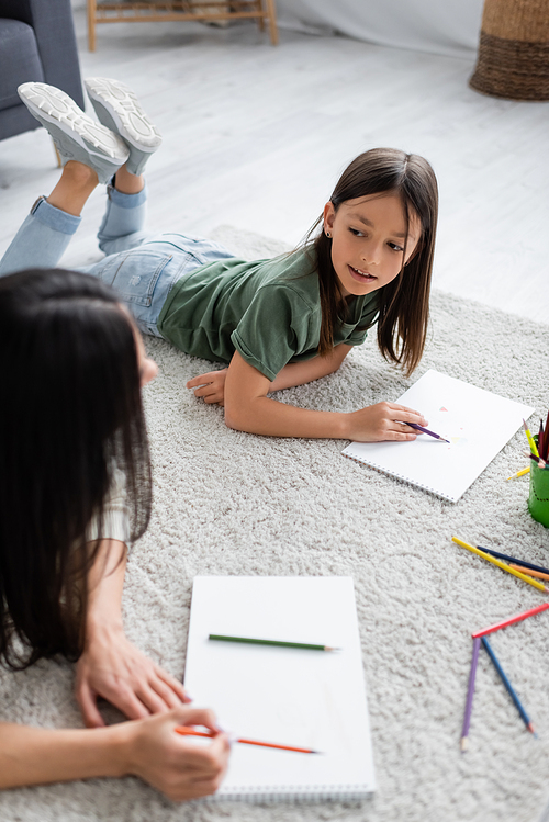 girl lying on carpet and holding pencil near paper while looking at blurred nanny