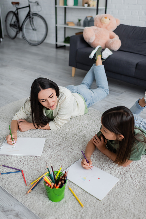 smiling babysitter drawing and looking at paper near girl in living room