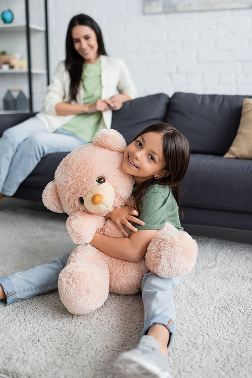 happy girl sitting on carpet and hugging teddy bear near blurred babysitter in living room