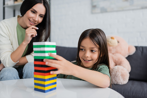 positive babysitter playing wooden tower game with pleased kid in living room