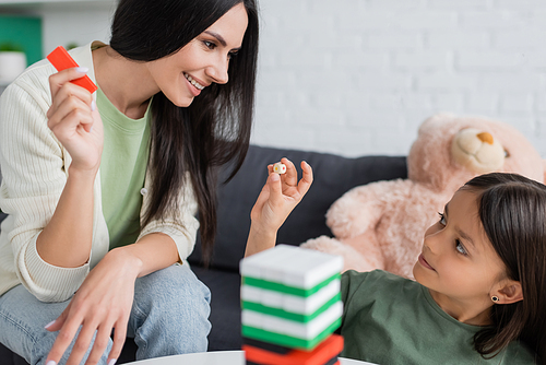 cheerful babysitter playing wood blocks game with girl in living room