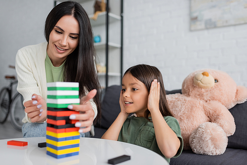 happy babysitter playing wood blocks game with smiling girl in living room