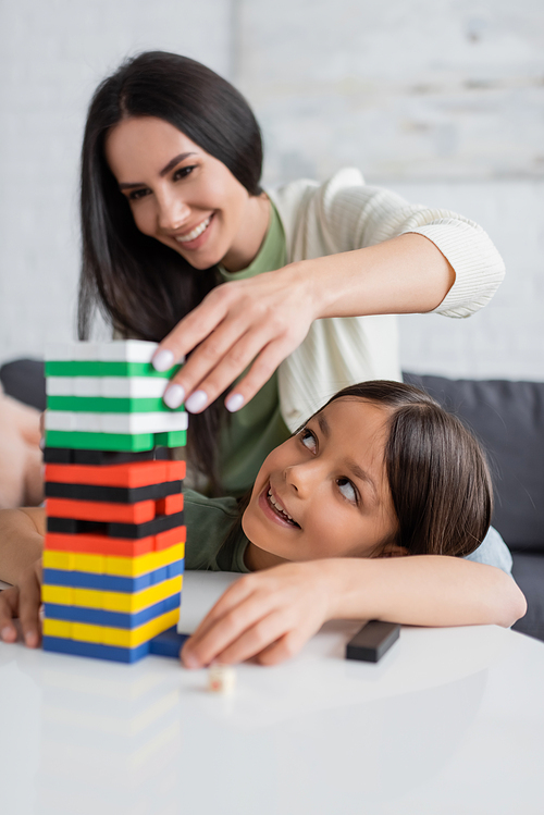 cheerful babysitter playing wood blocks game with pleased girl in living room