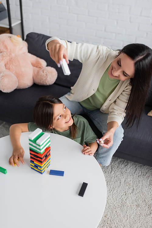 high angle view of positive babysitter playing wooden tower game with happy girl in living room