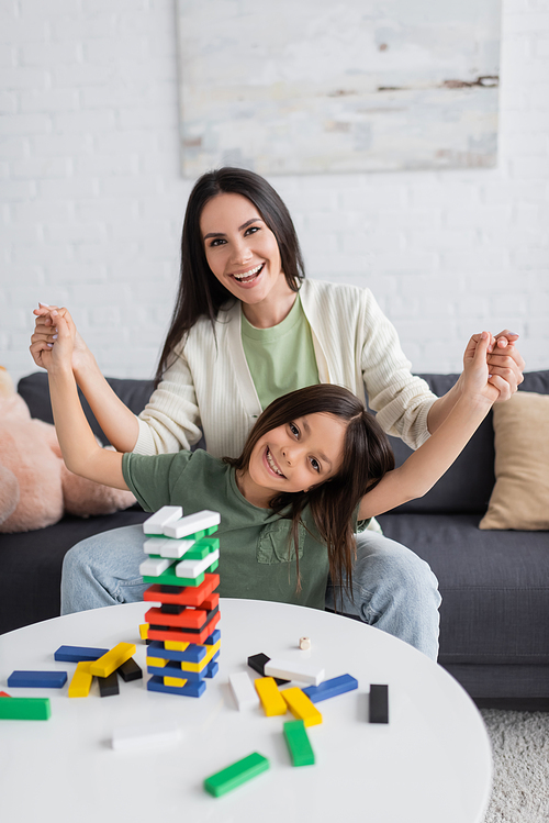 happy babysitter playing wooden tower game with joyful girl in living room