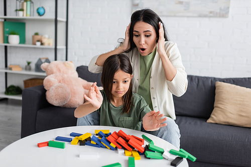 shocked nanny touching head near upset girl and colorful wooden blocks