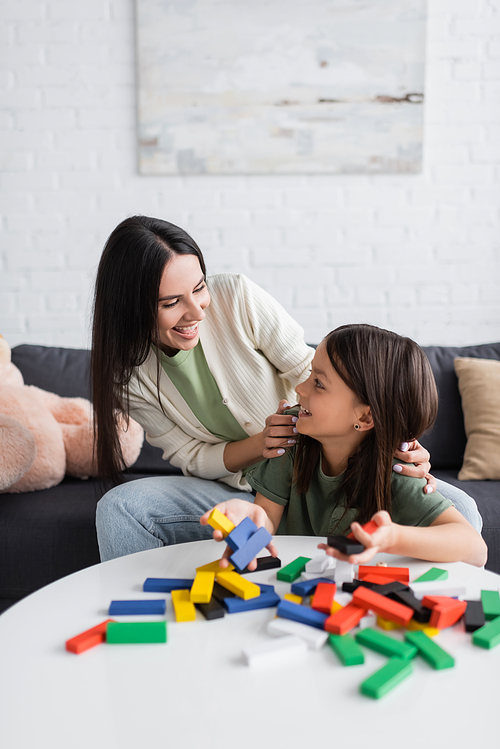 cheerful babysitter looking at girl near multicolored wooden blocks on table