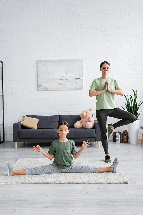 full length of nanny and girl with closed eyes meditating in yoga poses in living room