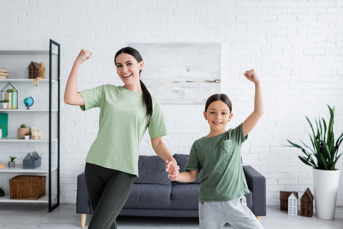 cheerful girl and babysitter holding hands and showing muscles in living room