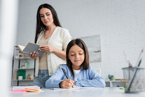 babysitter standing with book near girl writing in copybook at home