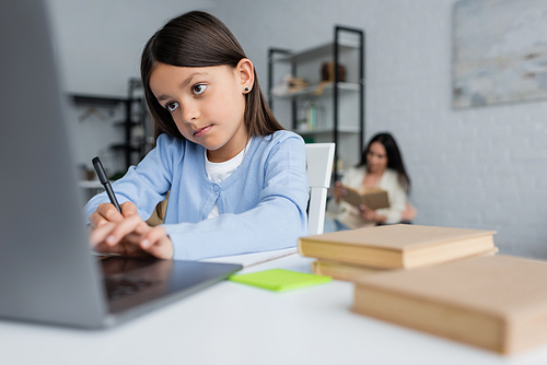girl writing near blurred laptop while doing homework near nanny on background