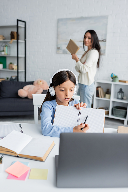 girl in headphones pointing at blank notebook near laptop and nanny on blurred background