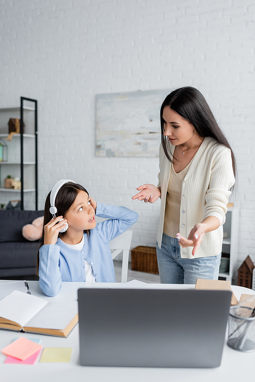nanny pointing at laptop near girl sitting in headphones during online lesson at home