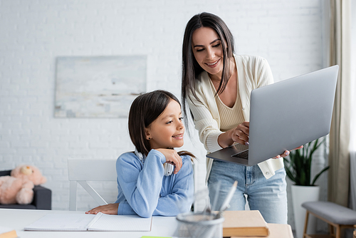 babysitter showing laptop to smiling girl doing homework on blurred foreground