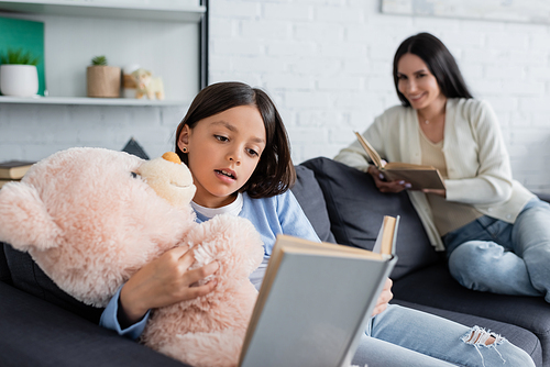 girl embracing teddy bear while reading book near nanny smiling on blurred background