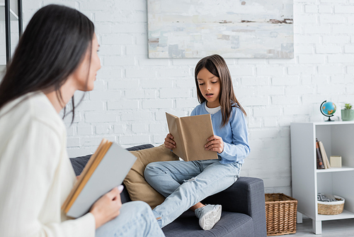 girl sitting on couch and reading book near blurred babysitter